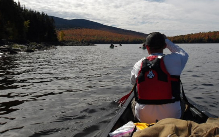 From the back of a canoe, you can see the person in front paddling. The boat is on calm water with trees lining the shore. 